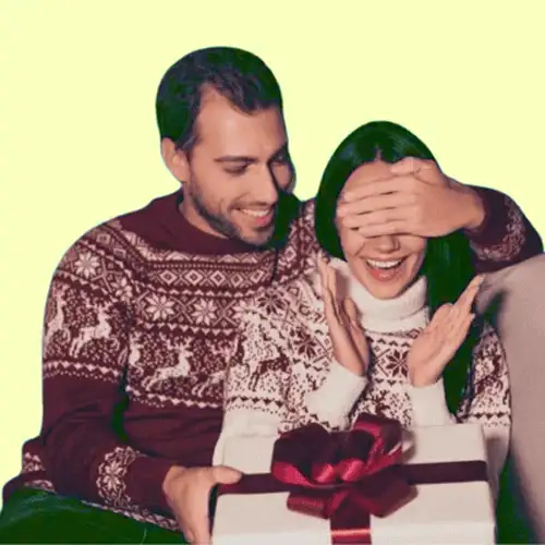 Couple in matching sweaters share a playful moment as the man covers the woman's eyes while presenting a gift with a red ribbon.