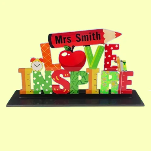 Colorful desk sculpture spelling 'LOVE INSPIRE' with an apple, clock, and pencil labeled 'Mrs Smith' on top.