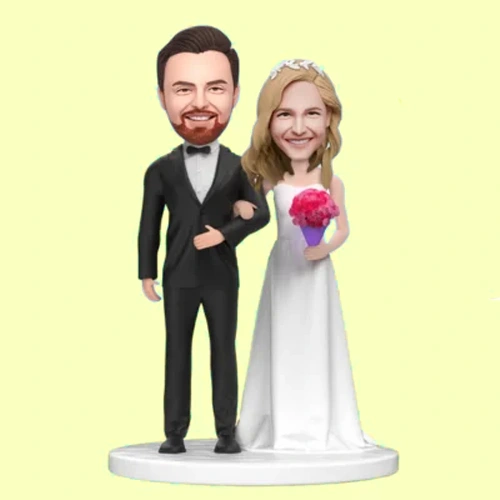 Wedding-themed figurine of a groom in a tuxedo and a bride in a white gown, holding a pink bouquet, standing together on a pedestal.
