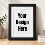 Framed white poster with bold text 'Your Design Here' on a wooden surface beside a plant and books.