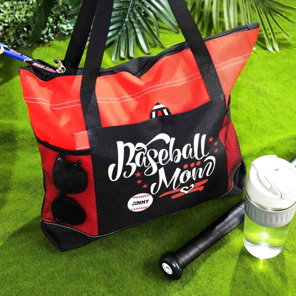 Red and black baseball bag with 'Baseball Mom' graphic on the side, a bat resting beside it on a green grass field, and a water bottle nearby.