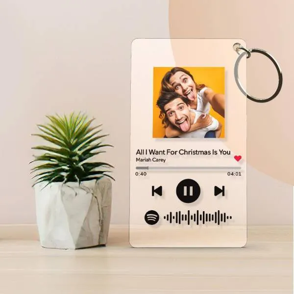 A smartphone-style music player mockup on a light desk beside a small potted plant, showing 'All I Want For Christmas Is You' by Mariah Carey with play/pause controls and a Spotify waveform, and a hanging key ring to the right.