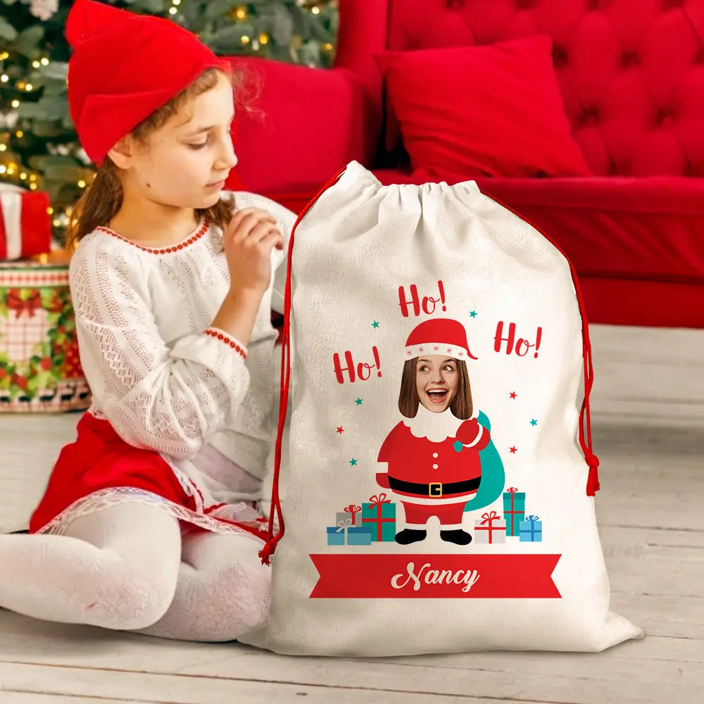 Young girl in a red Santa hat sits beside a large white drawstring gift bag with a cartoon Santa and a red 'Nancy' ribbon banner.