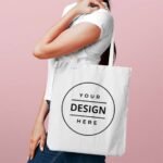 Person wearing a white shirt carries a white tote bag with a circular 'Your Design Here' logo against a pink background.