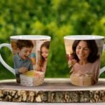 Two mugs printed with photos of smiling people, set on a wooden table outdoors with a green garden backdrop.
