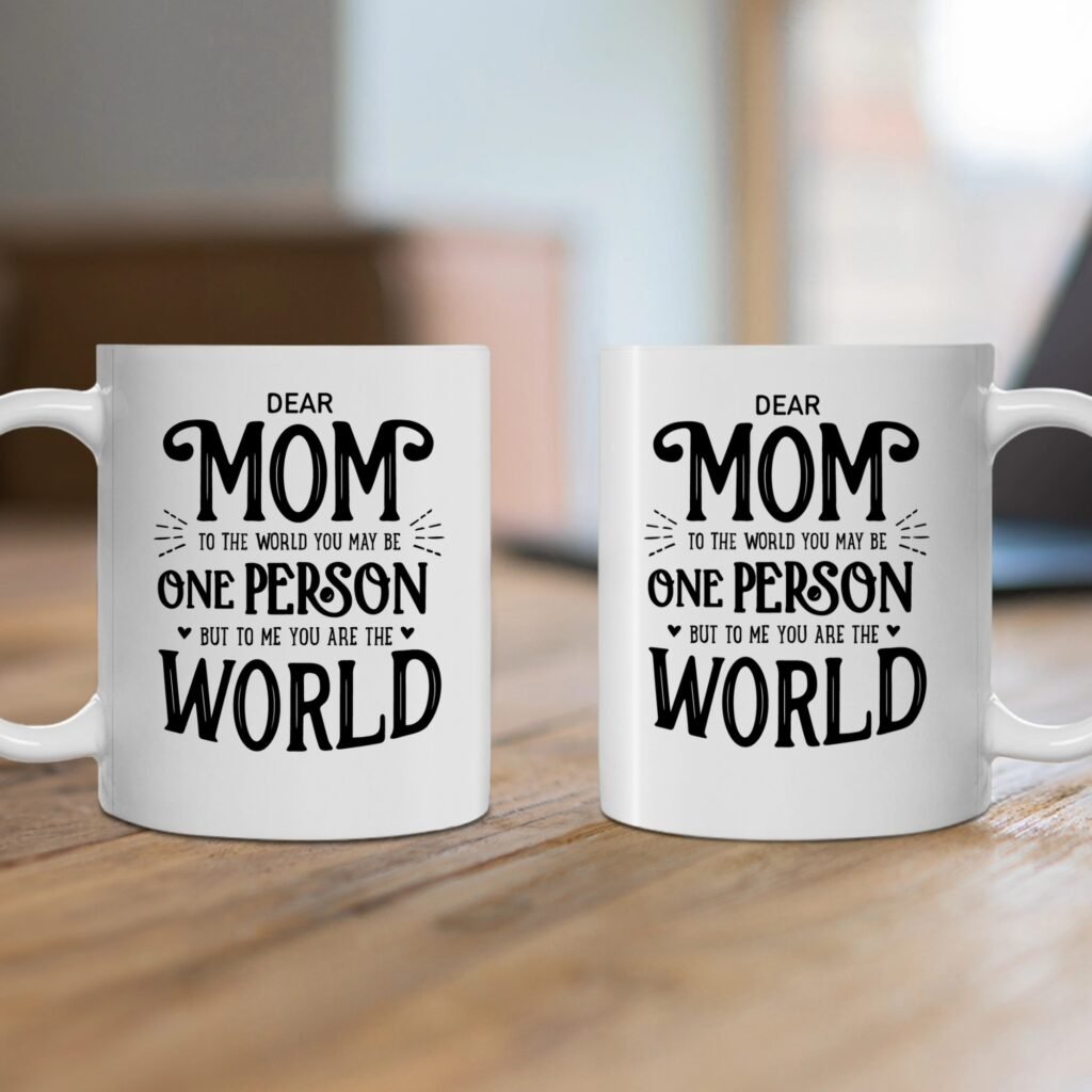 Two white mugs on a wooden surface display the message: 'Dear Mom, to the world you may be one person, but to me you are the world,' with a soft, blurred background.